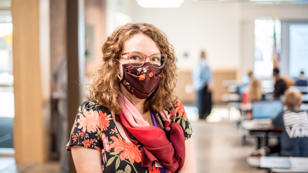 Student wearing a mask looking at the camera in the foreground with the rest of her class blurry in the background.