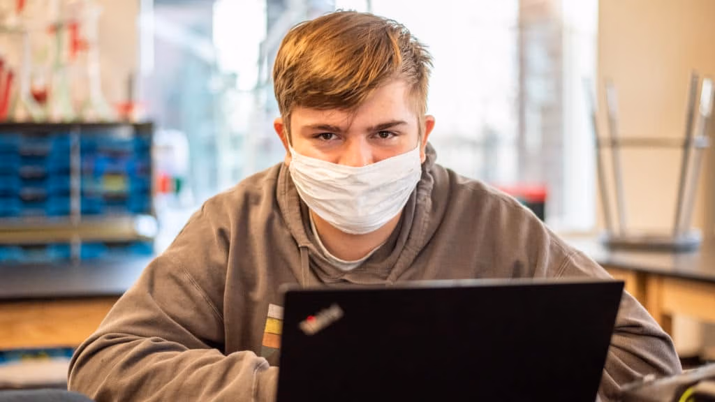 Student in a mask sitting a a desk in a classroom with their laptop in front of them.