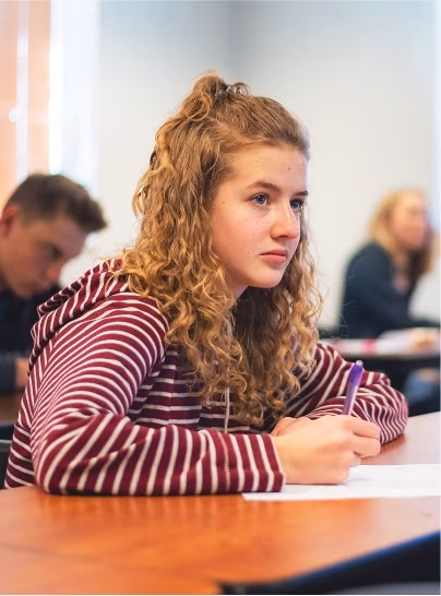 Female student in the foreground taking notes in school with other students in the background.
