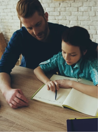 Father looking over his daughters shoulder helping her with homework at the dining room table.