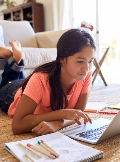 Female student laying on the floor in her home working on a laptop computer with papers and pens beside her.
