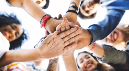 Bottom up image of a group of students with their hands in the middle stacked on top of each other.