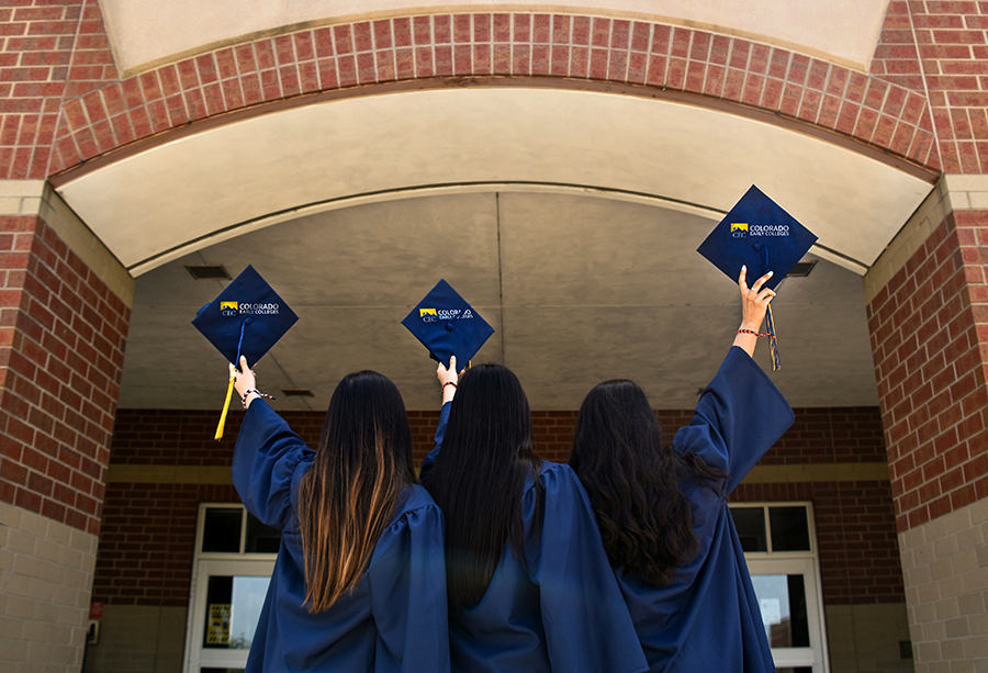 Three student graduates in gowns facing a school holding their caps up with one arm.