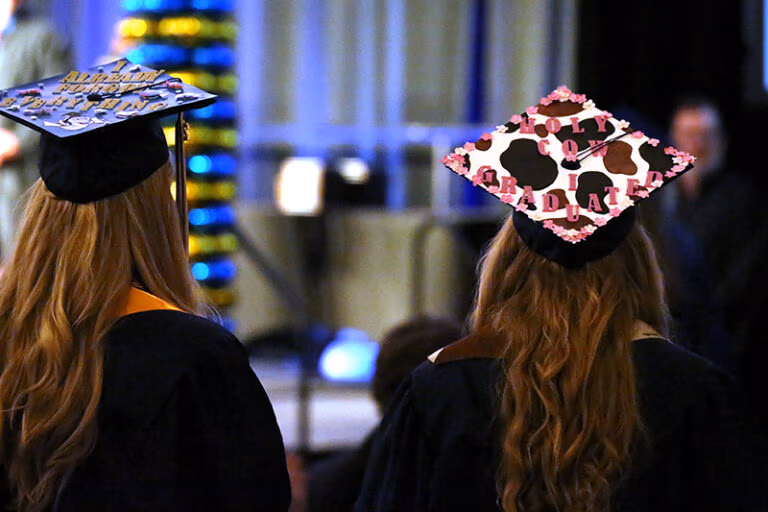 Two graduates from CEC Douglas County North wearing caps and gowns facing away from the camera and looking at the stage.