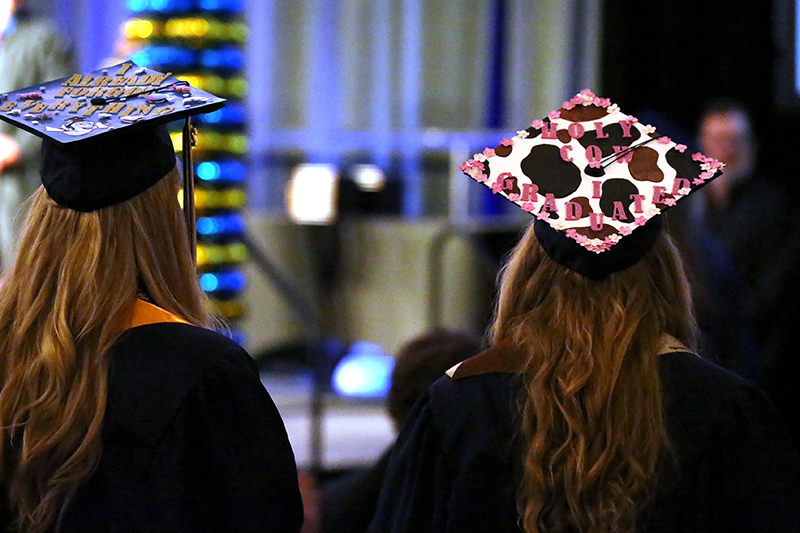 Two graduates from CEC Douglas County North wearing caps and gowns facing away from the camera and looking at the stage.