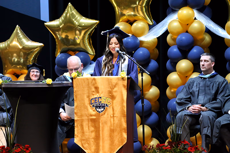 Leslie Mera Sanchez giving the senior class speech behind a podium with faculty on either side. There are yellow and blue balloon decorations.