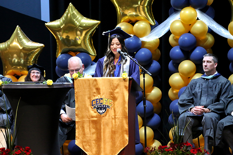 Leslie Mera Sanchez giving the senior class speech behind a podium with faculty on either side. There are yellow and blue balloon decorations.