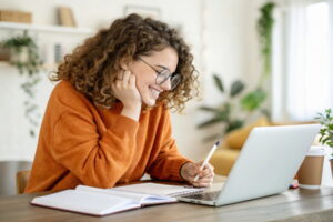 Smiling young woman with curly hair taking notes while studying online in a cozy home environment.
