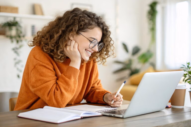 Smiling young woman with curly hair taking notes while studying online in a cozy home environment.