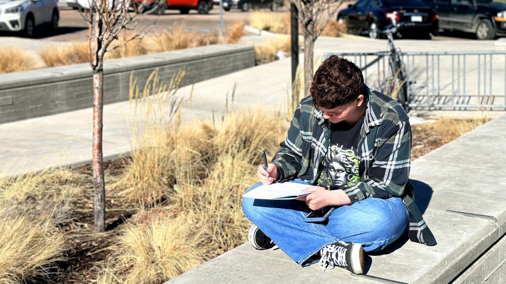 CEC Colorado Springs Student sitting outside on a ledge writing in a notebook.