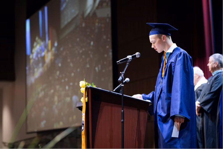 CEC Fort Collins High School graduation speaker standing at the podium giving a speech. Wearing a blue cap and gown.
