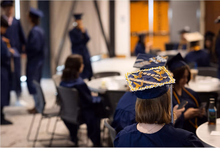 Top of a graduation cap in focus in the foreground with other graduates blurred in the background. The cap has butterflies, flowers, and scrabble tiles.
