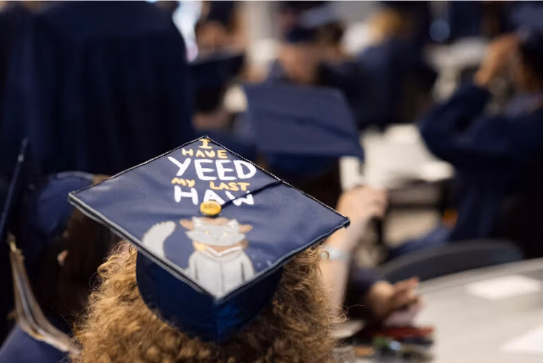 Top of a graduation cap in focus in the foreground with other graduates blurred in the background. The cap says, "I have yeed my last haw" with a picture of a cat.