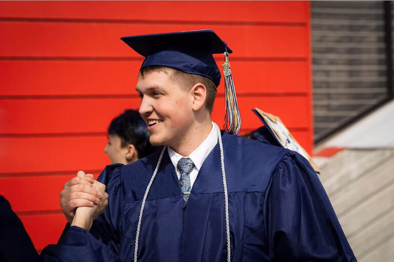 CEC Fort Collins High School graduate claps hands with another graduate outside after graduation. There is a bright red wall in the background.