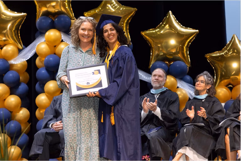 2025 CECFC Graduate receives an award on stage from a faculty member with other faculty in the background clapping.