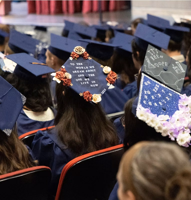 Image of the back of CECFC graduates seated at the 2025 graduation ceremony. The cap in focus says, to the world we dream about and the one we live in now, written on top of it.
