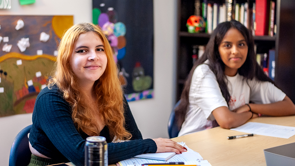 Two students sitting at a desk in a classroom looking at the camera and smiling.