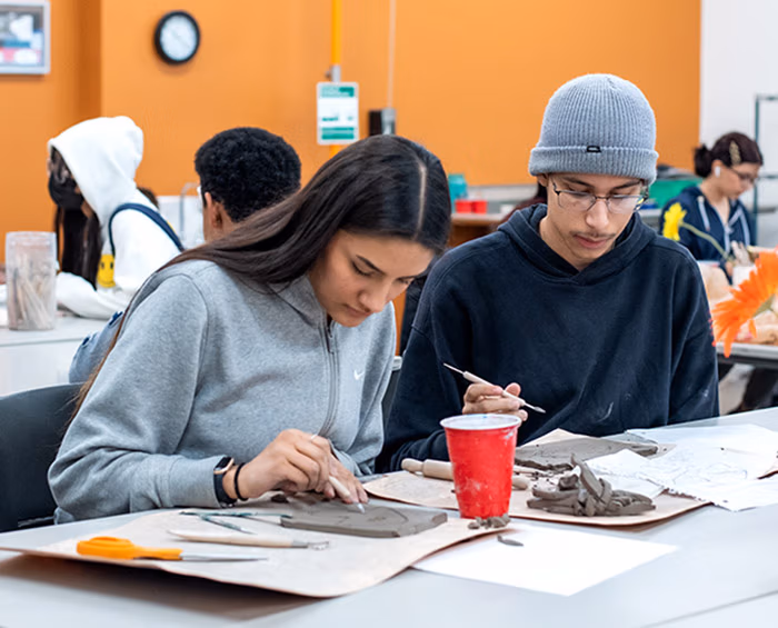 Two students sitting at a table in a classroom working on a sculpture project.