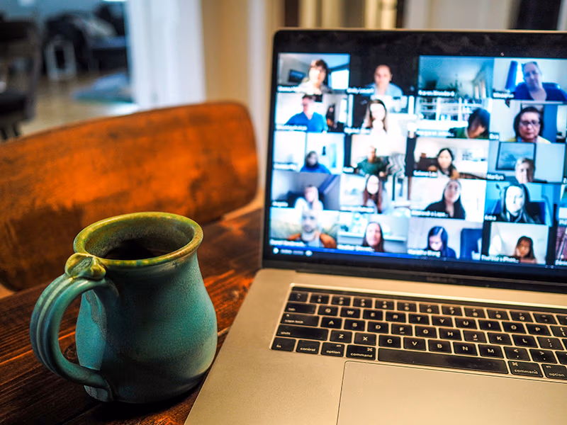 Laptop with images of people in an online meeting. The laptop is sitting on a table with a cup of coffee sitting next to it.