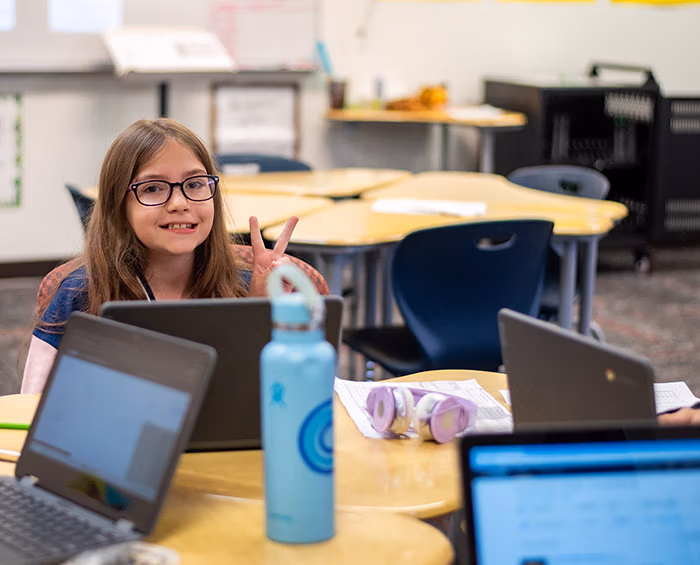 CECCS Student giving a peace sign while sitting at a desk with a computer in front of her.