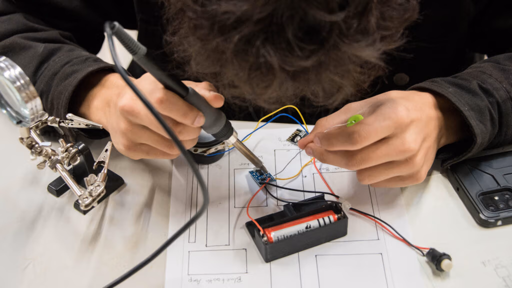 CEC Student soldering a computer chip with wires that lead to a battery.