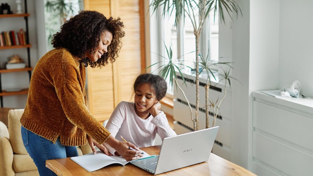 A mother works with her daughter on homeschool work at a table with a laptop and a notebook.