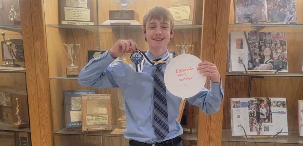 Ron Baravick holds up first place DECA medal and a paper plate that says, "Colorado Early College 1". Ron is standing in front of a trophy case.
