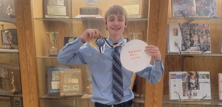 Ron Baravick holds up first place DECA medal and a paper plate that says, "Colorado Early College 1". Ron is standing in front of a trophy case.