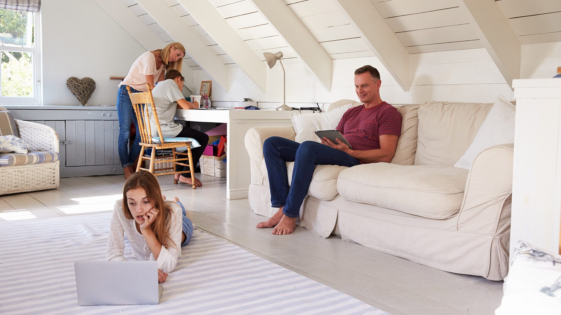 Family in a room, Dad is sitting on the couch with a tablet, the daughter is on the floor looking at a computer doing homework, mom and son are sitting at a desk working on school work.